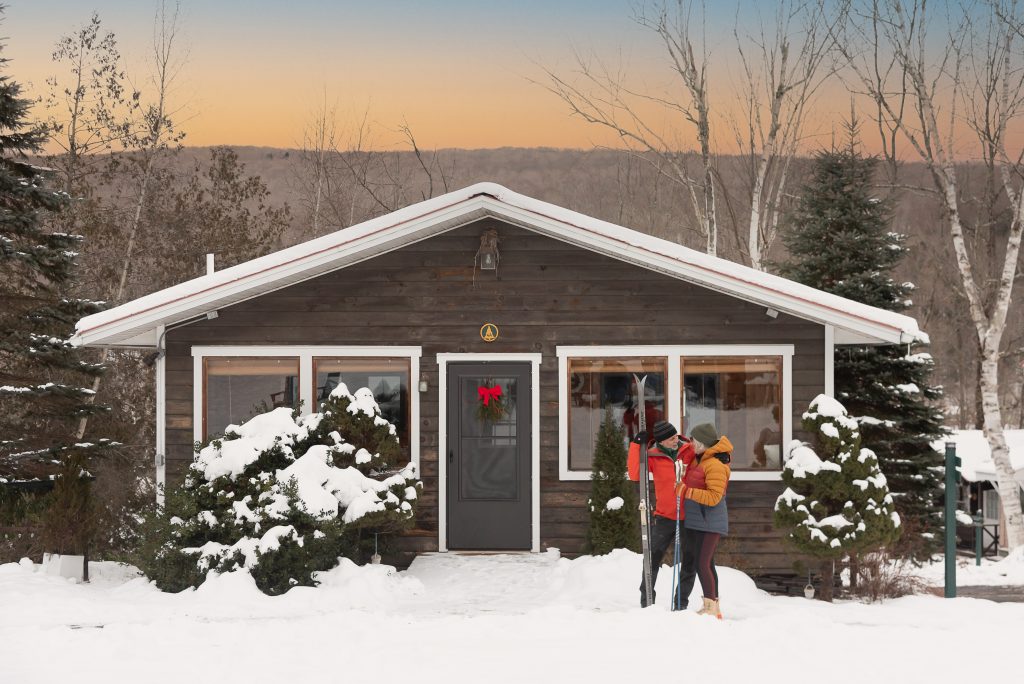 Cozy brown cabin decorated with a holiday wreath, snow-covered trees, and two people with skis in front at sunset.