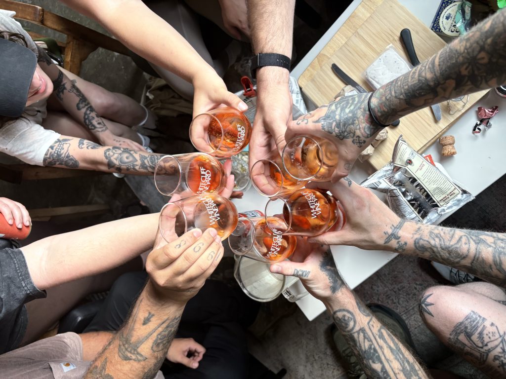 A group of people with tattooed arms raising wine glasses together in a toast over a table with snacks and cheese, viewed from above.