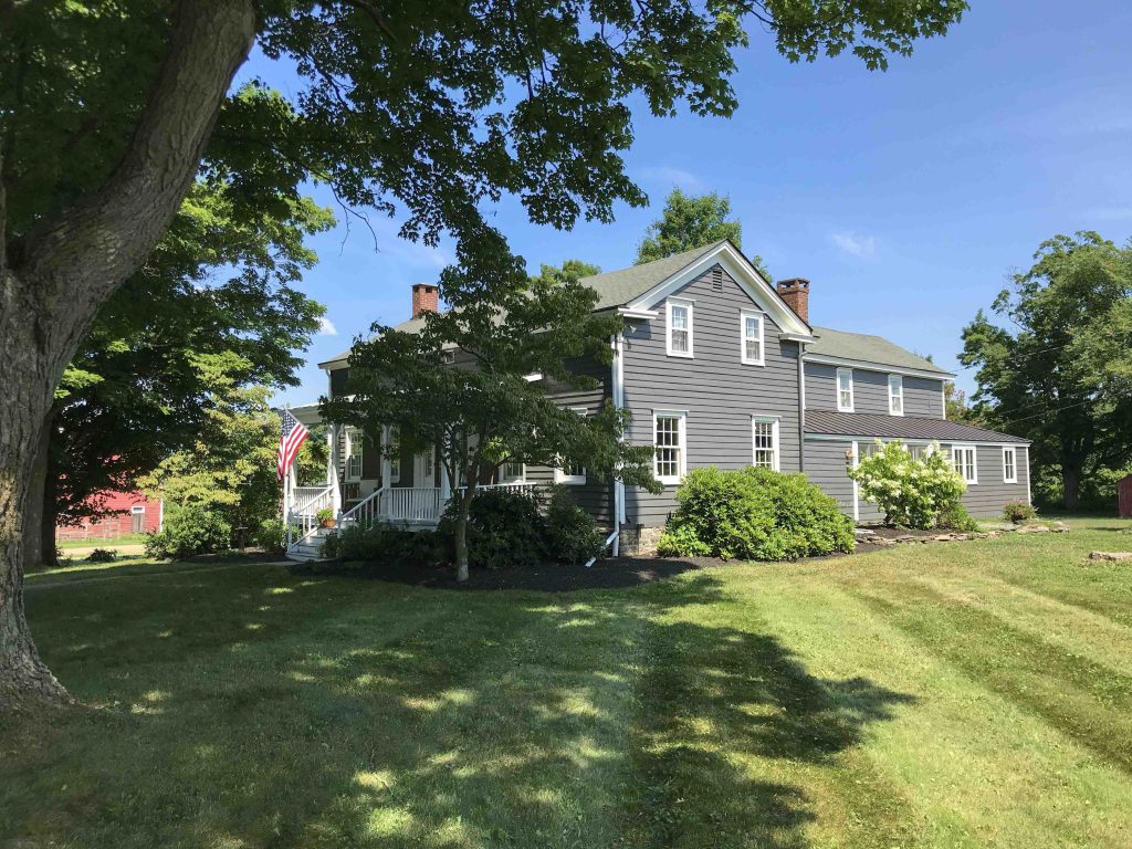 Gray two-story house with white trim, front porch, and American flag, surrounded by green lawn and trees on a sunny day.