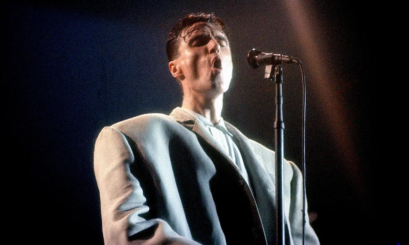 David Byrne in a large suit singing at a microphone