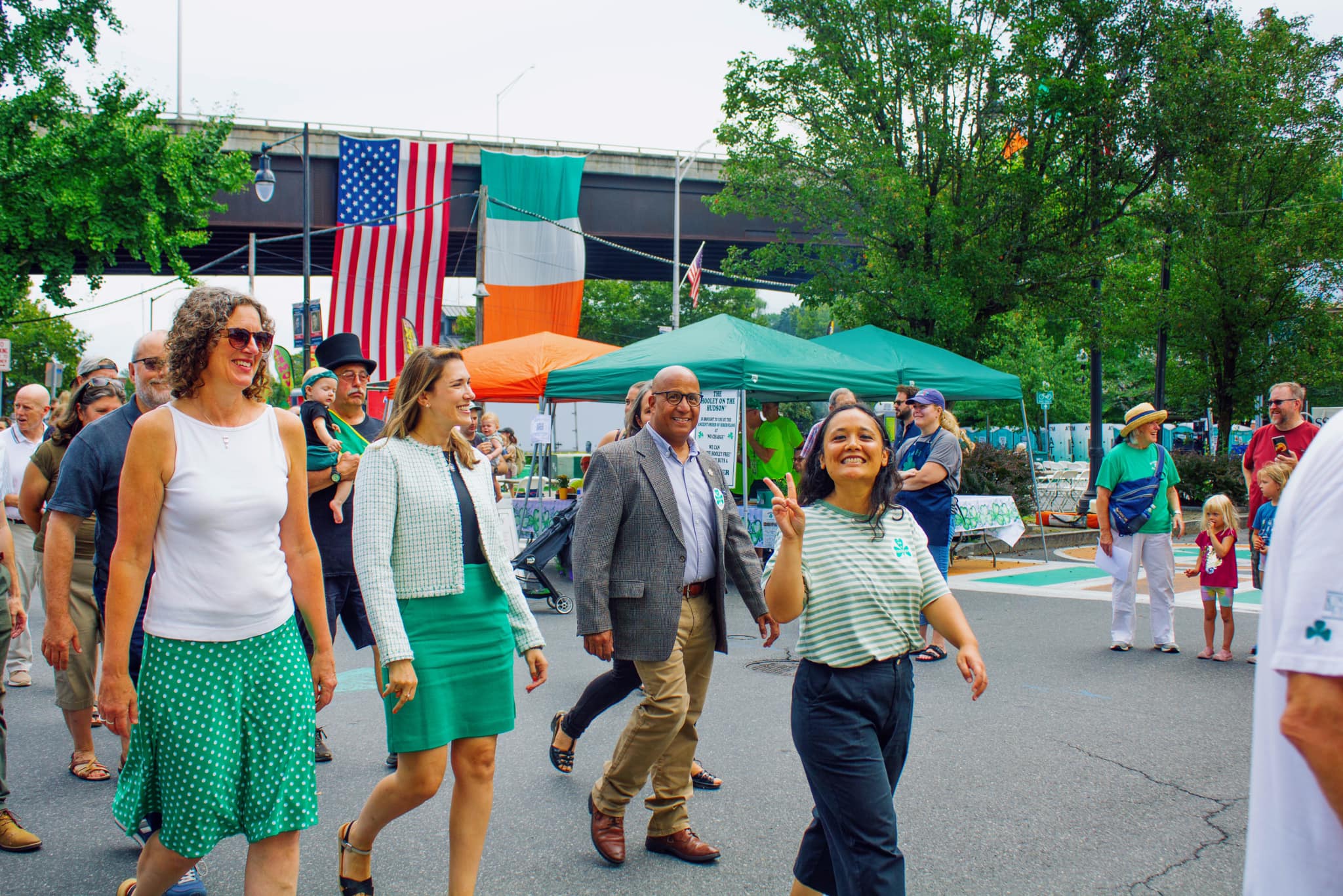 People walk at outdoor festival with American and Irish flags, green tents and trees, adults and children enjoying the event on a street.