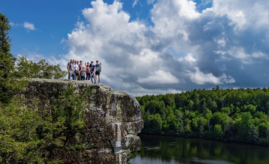 Nine people stand together on a rocky cliff, overlooking a forest and lake under dramatic clouds.