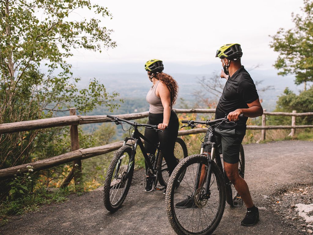 Two cyclists wearing yellow helmets pause with their bikes at a wooden fence, looking out over a scenic vista with trees and distant hills.