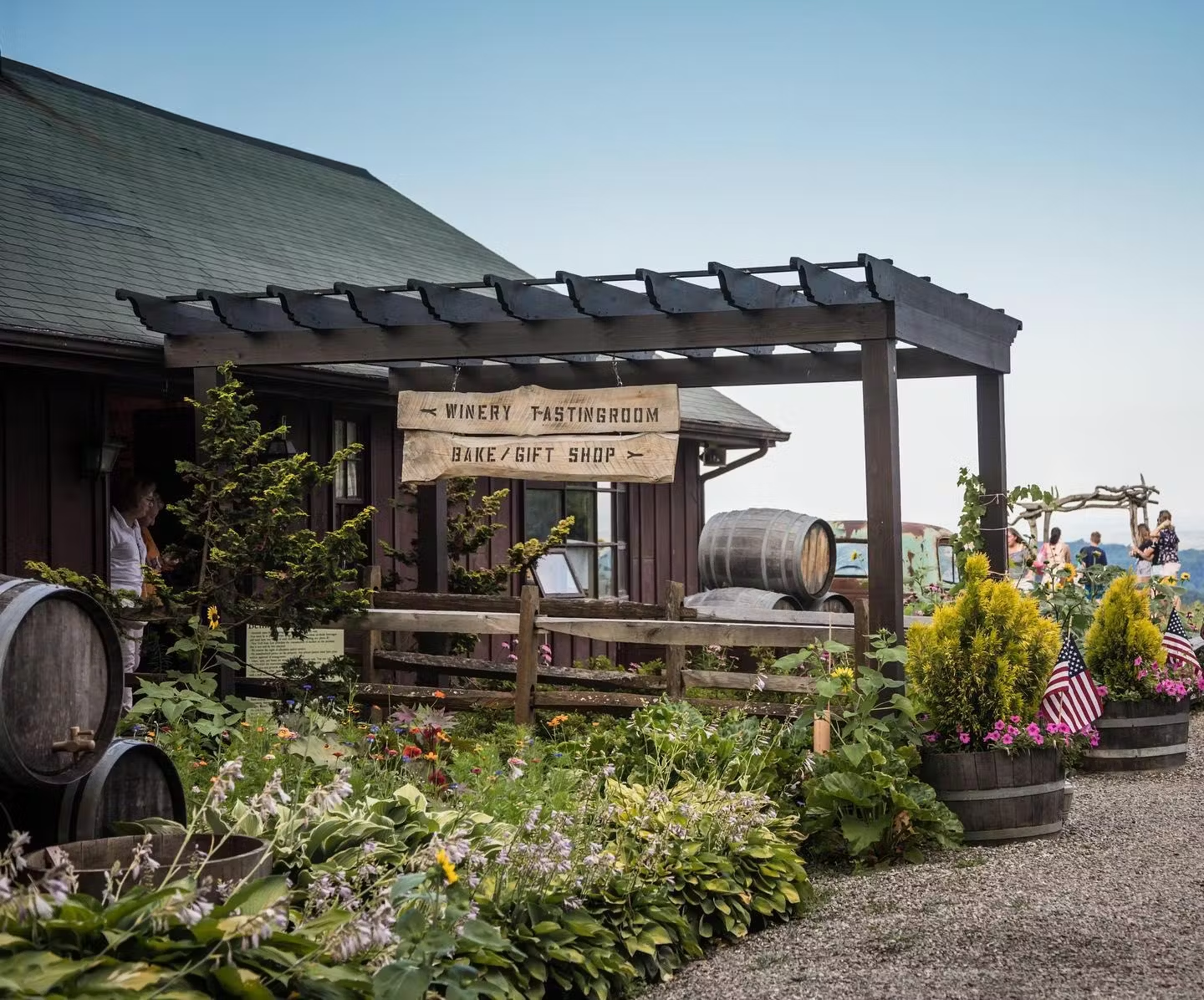 Winery tasting room and bake shop entrance with rustic pergola, colorful flowers, barrels, American flags. People enjoy the outdoor scene.
