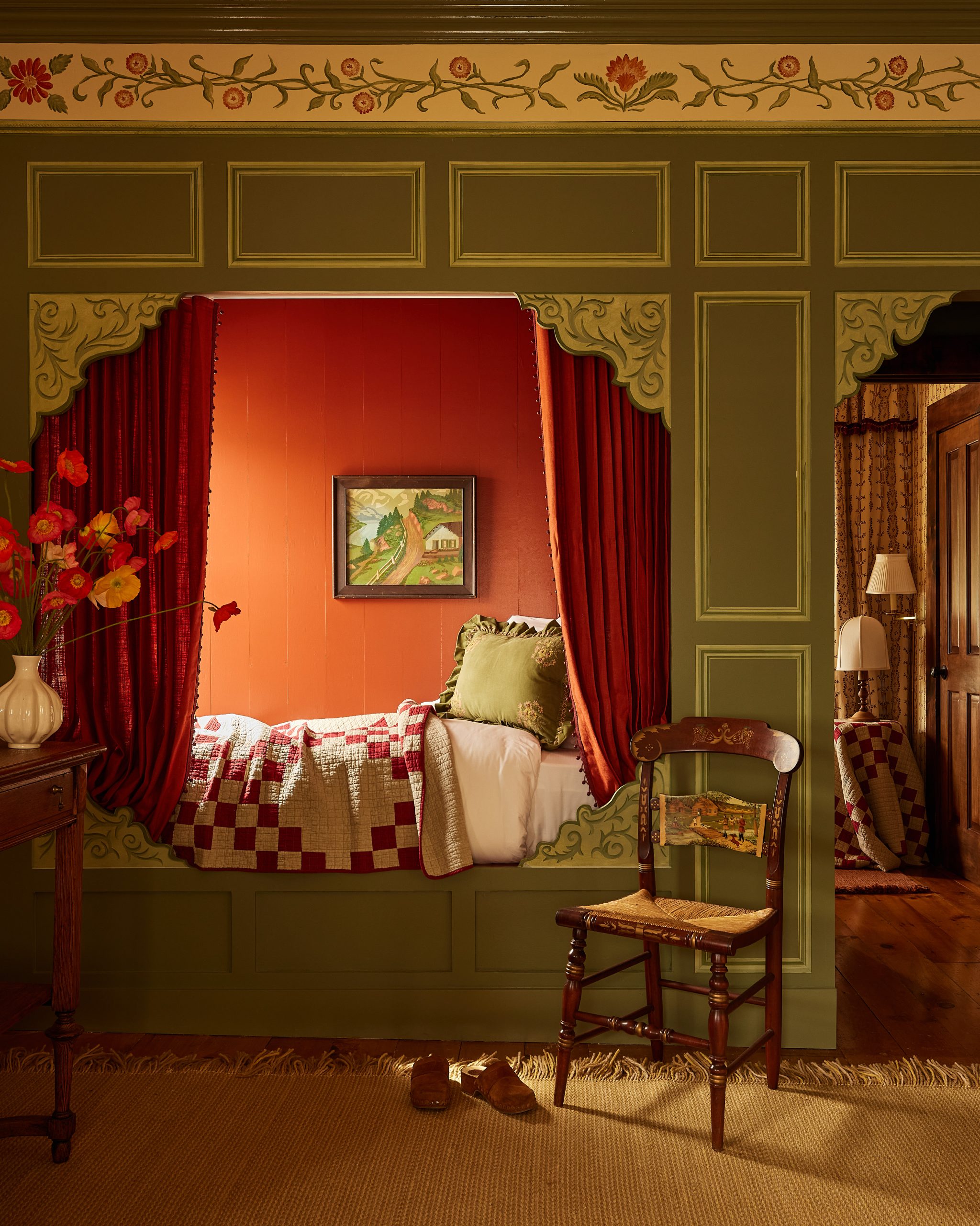 Cozy nook bed with red curtains, patterned quilt, green floral-painted woodwork, plus a chair and wooden slippers on the rug.