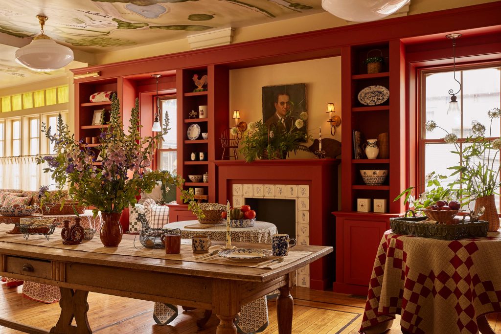 Warm dining room with red cabinets, rustic table, floral centerpiece, decorative plates, and painted ceiling.