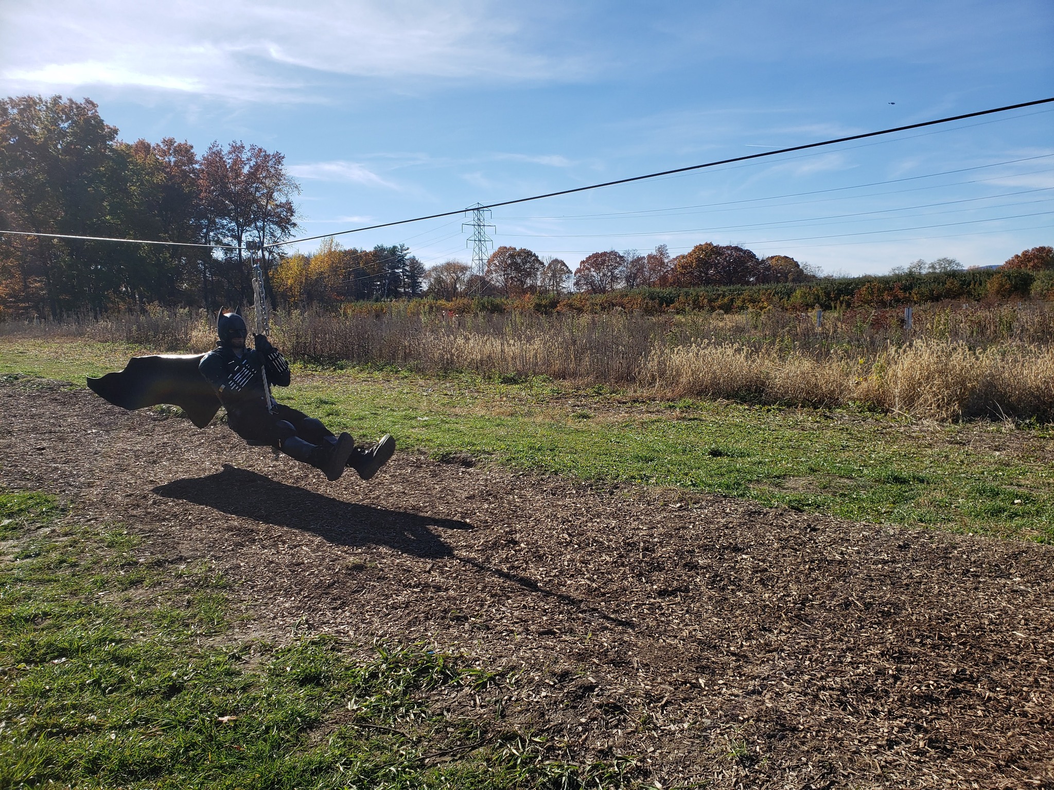 Person dressed as Batman riding a zipline over a grassy field at Hurds Family Farm during Spooktacular Weekend.