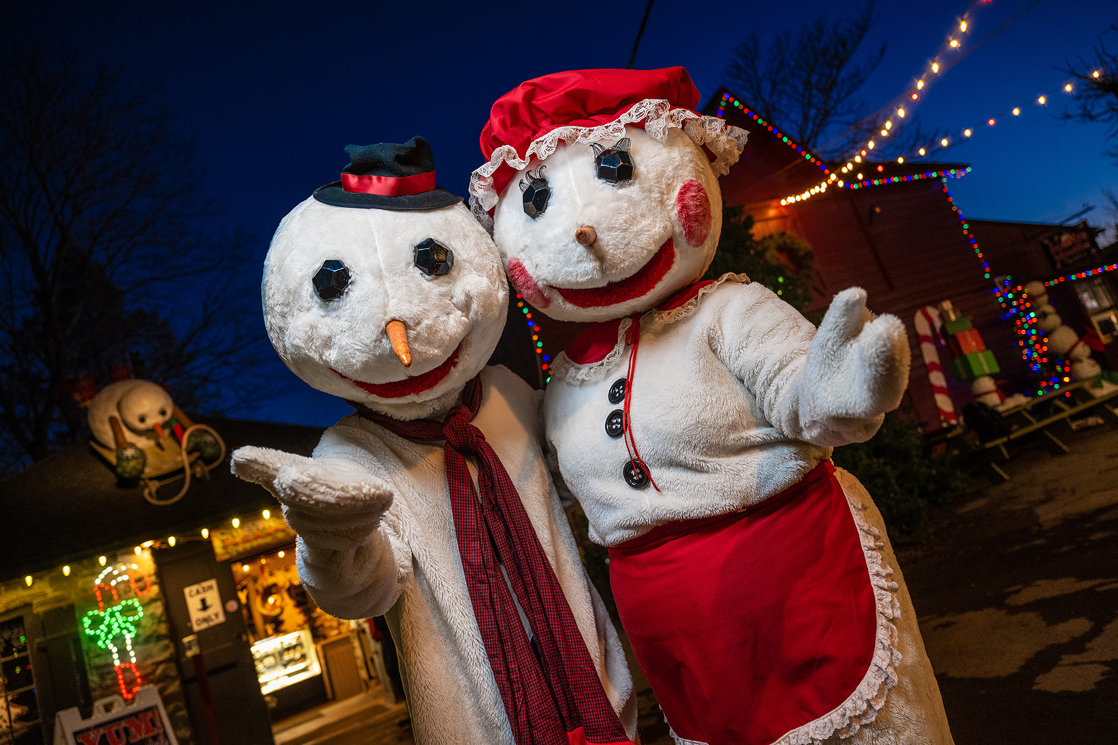 Two people in snowman costumes, one with red scarf and the other with red bonnet and apron, at festive outdoor holiday event with lights.