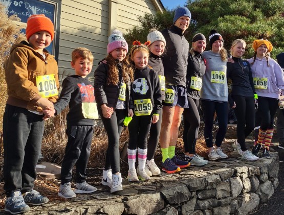 Group of adults and children wearing race bibs and winter hats, smiling on a stone wall at the Family of New Paltz Turkey Trot.