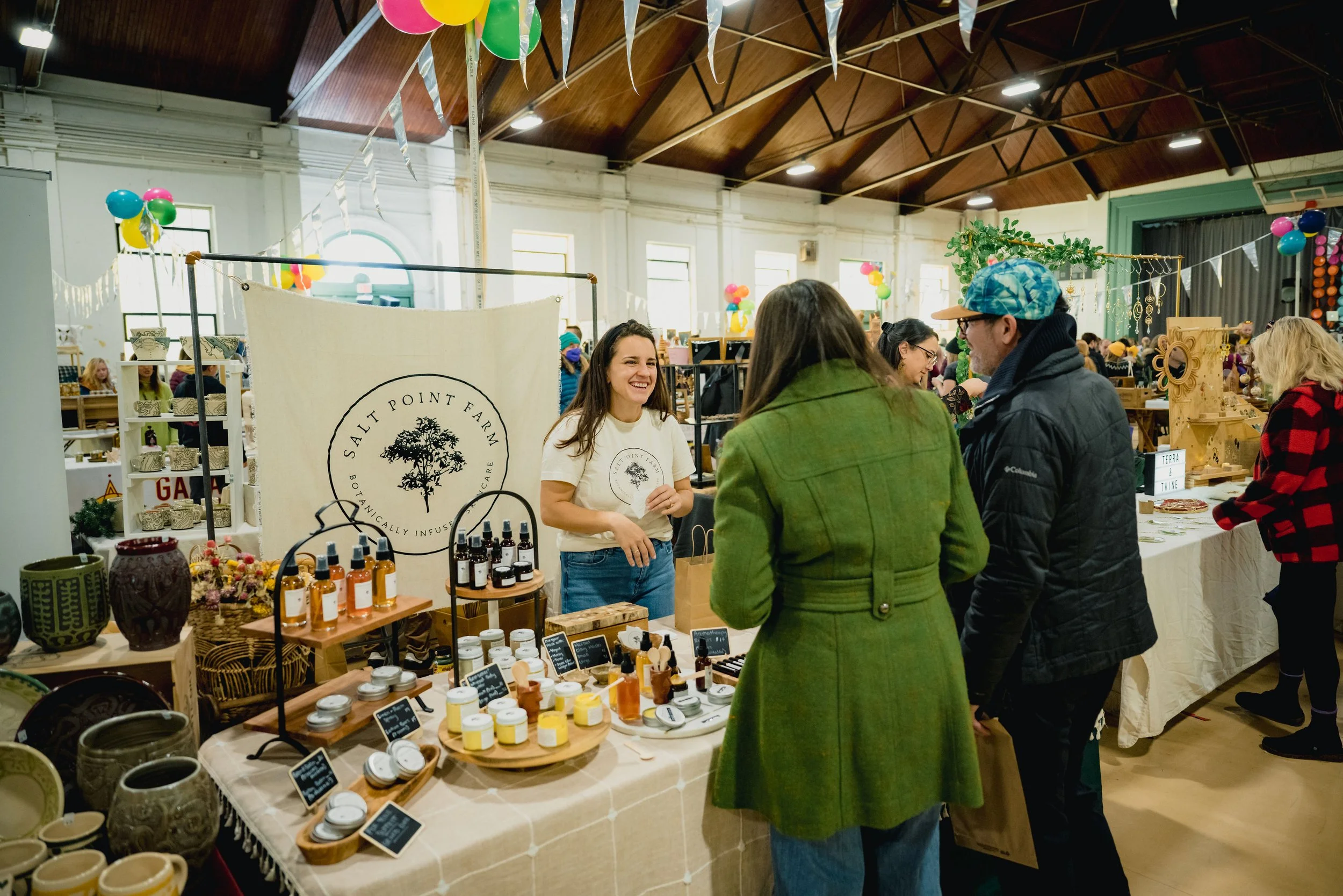 Vendor with Salt Point Farm banner smiles and talks with shoppers at holiday market booth displaying artisan goods and homemade products.