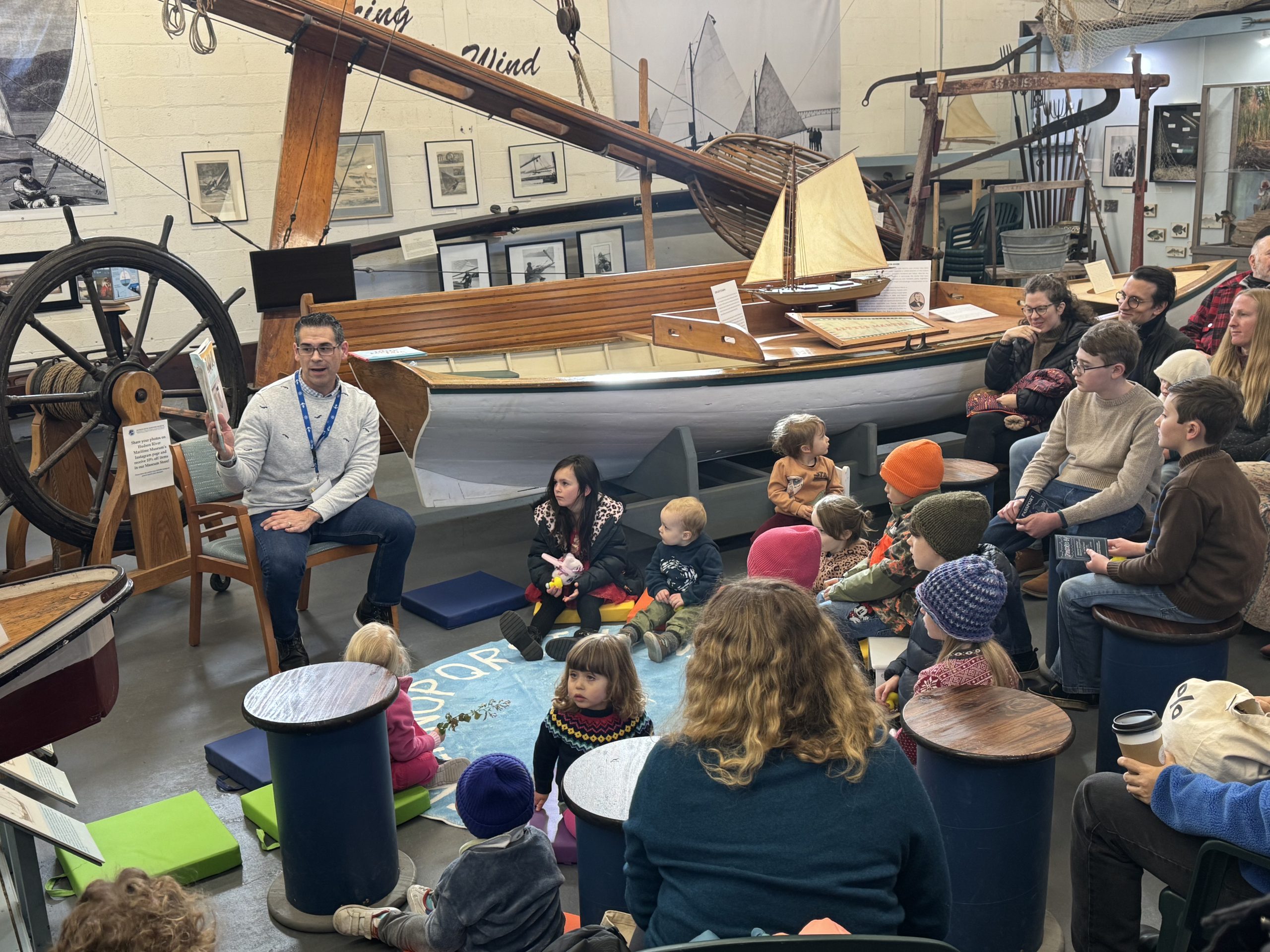 Man reading to children and adults inside a boat museum, with sailboats, oars, and nautical exhibits around them.