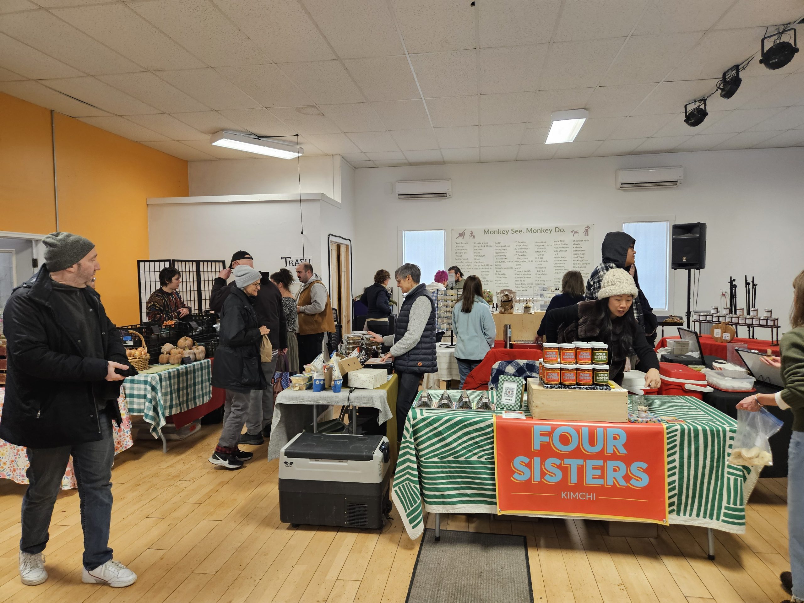 Indoor farmers market with people browsing vendor tables of produce, food, and kimchi under fluorescent lights.