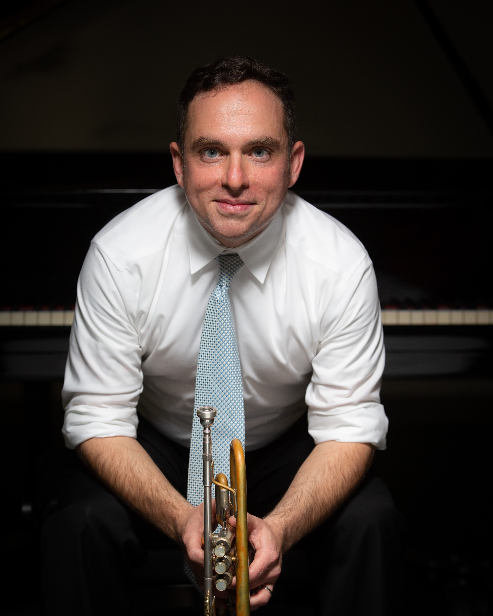Man in a white shirt and light blue tie seated in front of a piano, holding a trumpet.