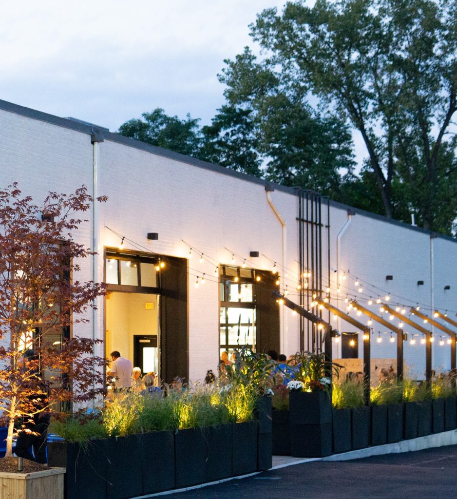 Evening patio seating at Blue Duck Brewing, with diners under string lights beside a white building and planters.