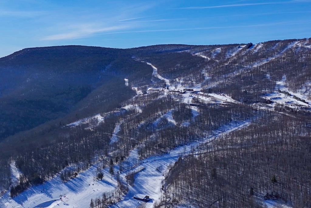 Aerial view of snow-covered ski trails winding through a forested mountain landscape under a clear blue sky.