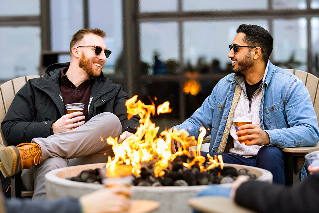 Two people sitting by a fire pit outdoors, holding drinks and talking with others around them