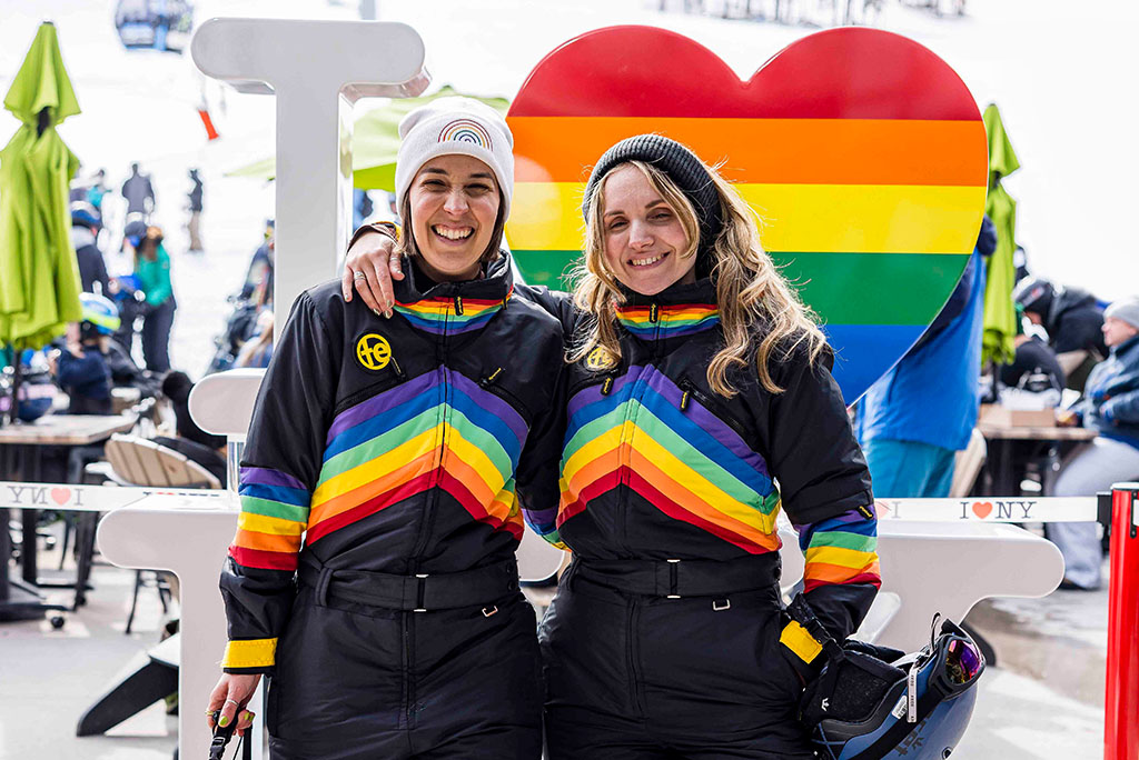 Two people in matching rainbow-patterned ski suits stand arm in arm in front of an ‘I ♥ NY’ sculpture with a rainbow heart at a ski resort.