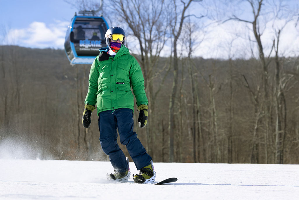 Person snowboarding on a sunny day, wearing a green jacket and helmet, with a ski lift gondola and bare trees in the background.
