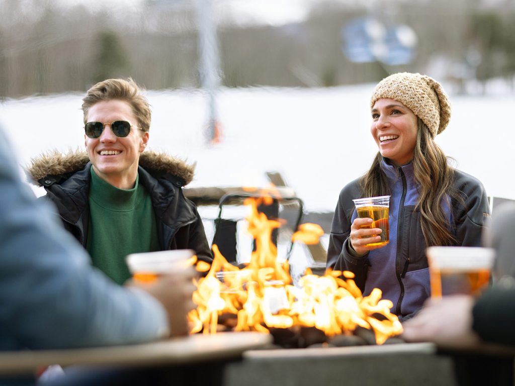 People gathered around an outdoor fire pit, holding drinks and wearing winter jackets and hats, with snow in the background.