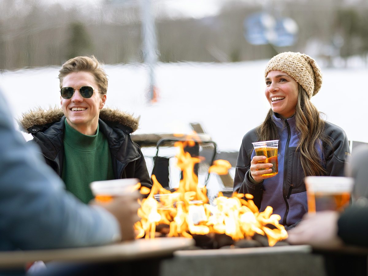 People gathered around an outdoor fire pit, holding drinks and wearing winter jackets and hats, with snow in the background.