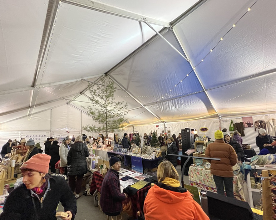 Indoor holiday market under a tent with shoppers browsing craft and gift vendors, festive lights, and seasonal decorations.