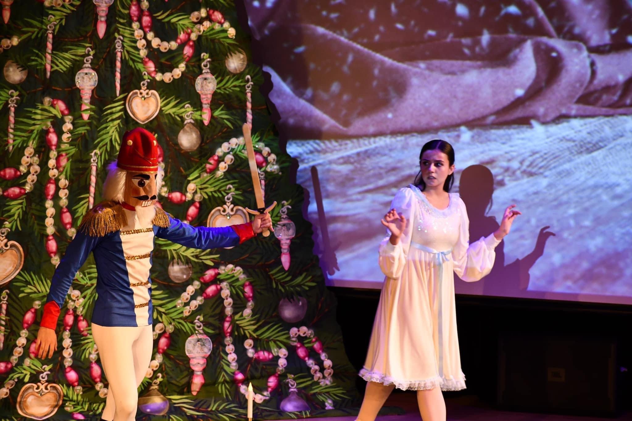Nutcracker performer in blue and white costume with sword stands beside dancer in white dress, in front of a decorated Christmas tree on stage.
